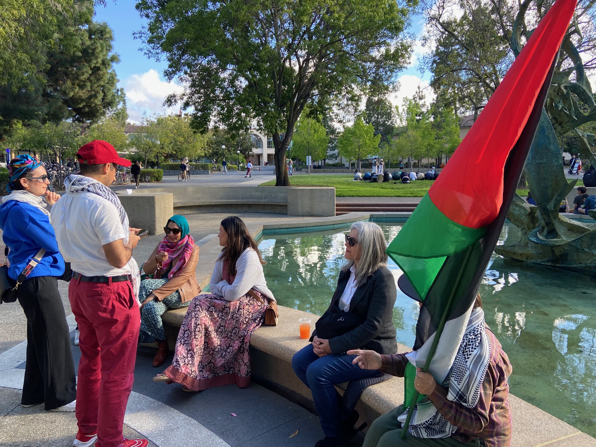 6 people rest on wall of fountain, one carries a Palestinian flag they are chatting